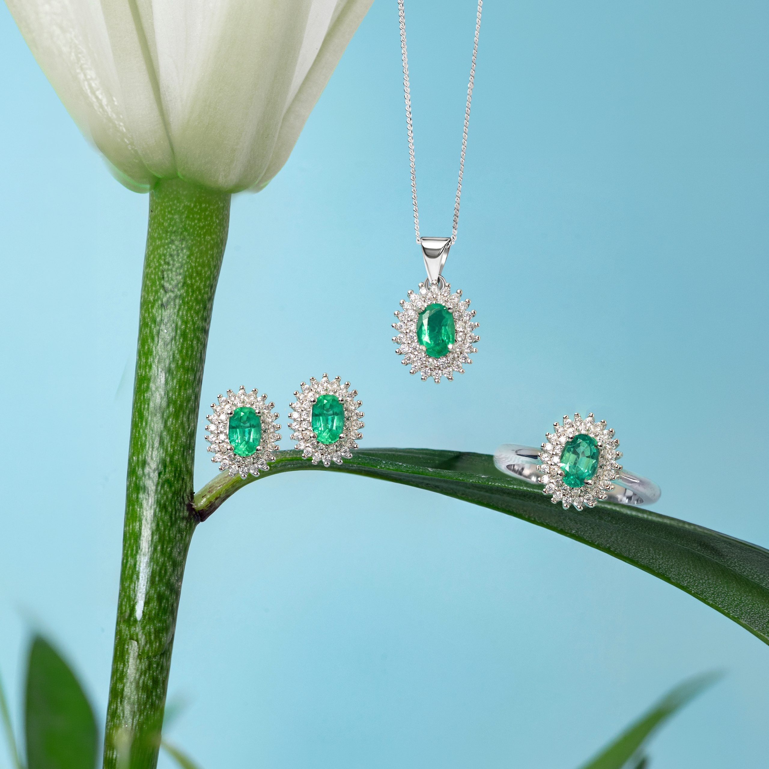 _NZR1333 Silver necklace with an emerald pendant surrounded by diamonds hanging from a green plant stem against a blue background.